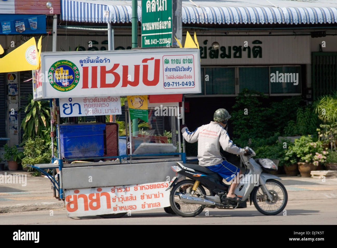 A vendor is pulling a food cart while riding a motorcycle on a city street in Khon Kaen, Thailand. Stock Photo
