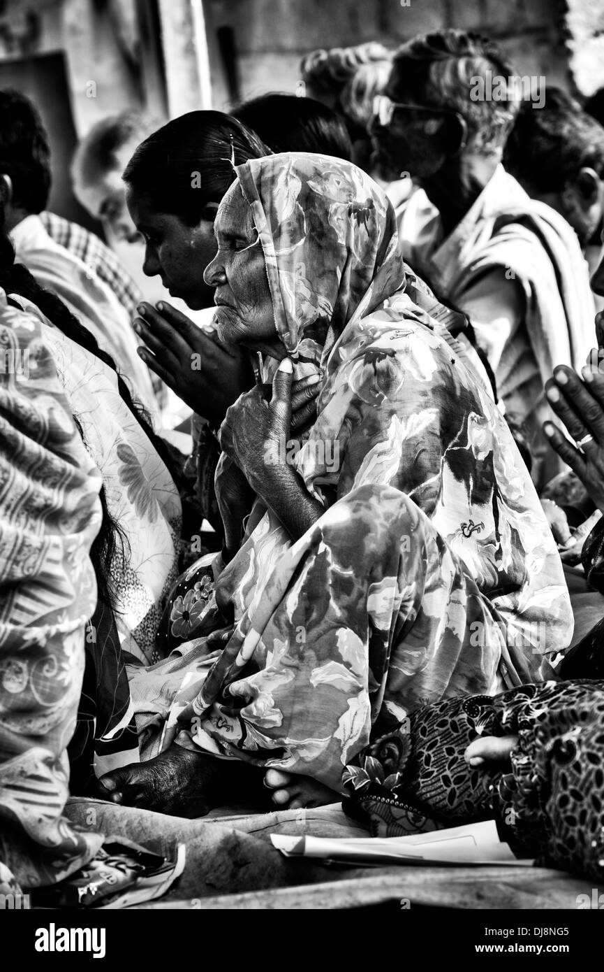 Old Indian woman in prayer whilst waiting at Sri Sathya Sai Baba mobile outreach hospital. Andhra Pradesh, India. Monochrome Stock Photo