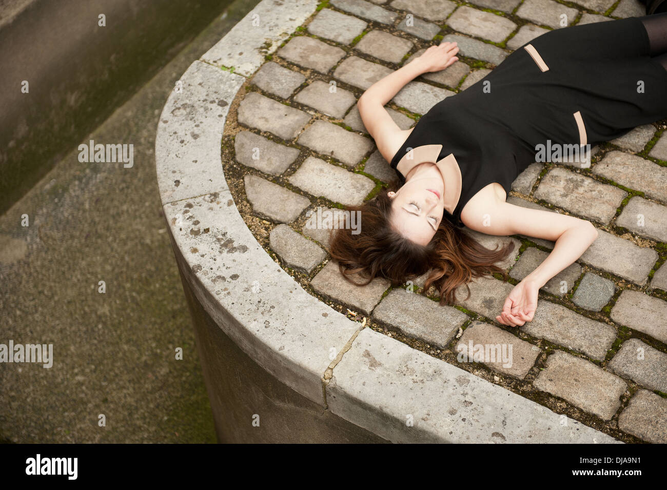 Elevated view of a woman wearing a black dress and lying down (dead?) on a cobbled road. Stock Photo