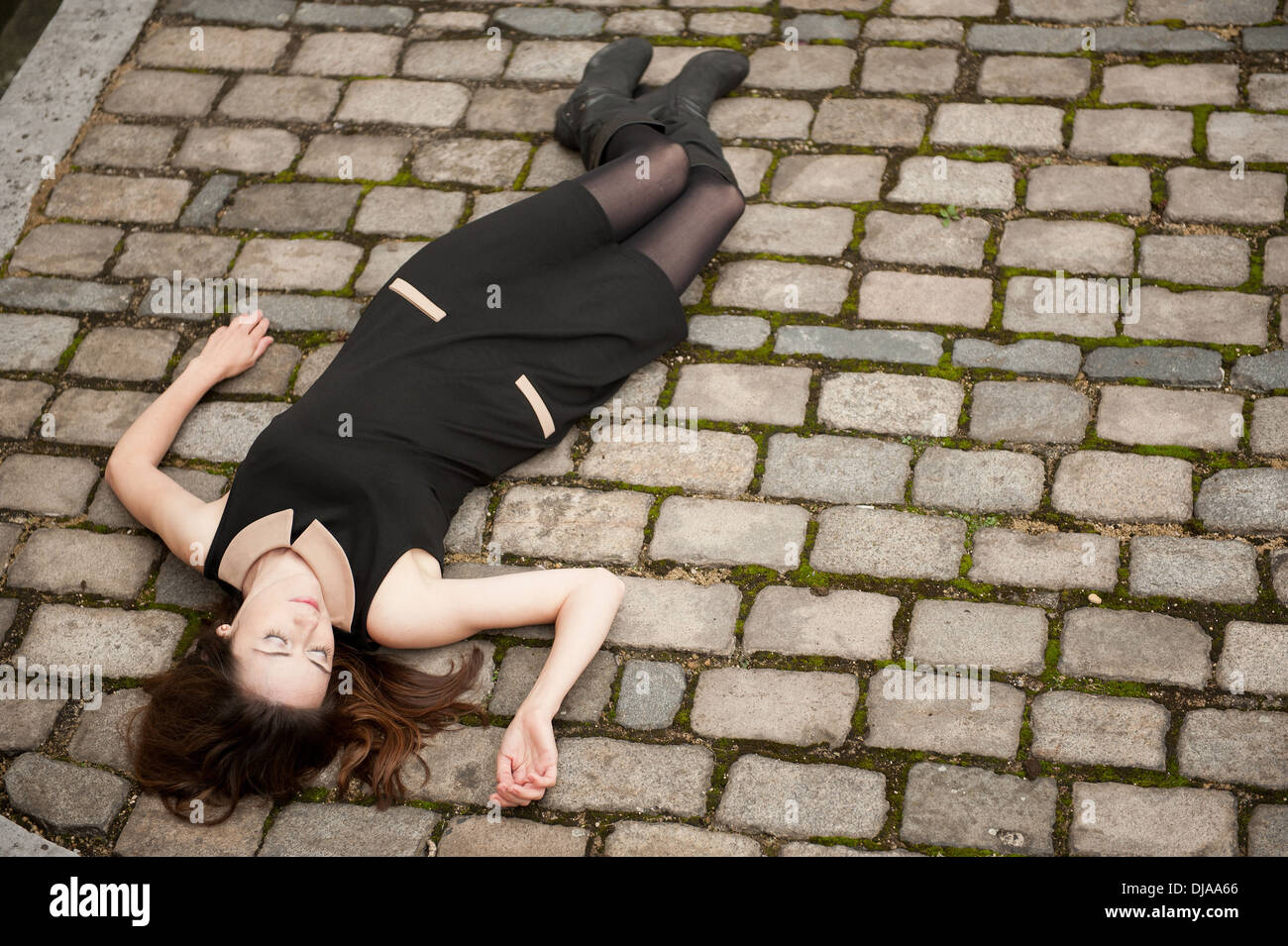Elevated view of a woman wearing a black dress and lying down (dead?) on a cobbled road. Stock Photo