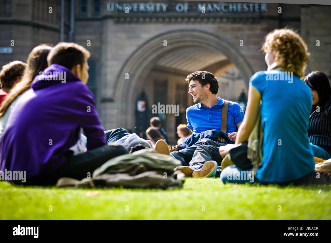group of multi racial Manchester university students sat on grass on campus in sunshine talking and laughing looking happy Stock Photo