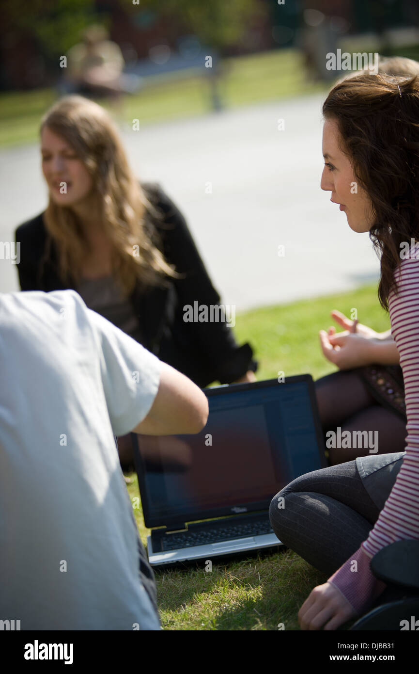 female university students sat on grass with laptop computer talking in sunshine Stock Photo
