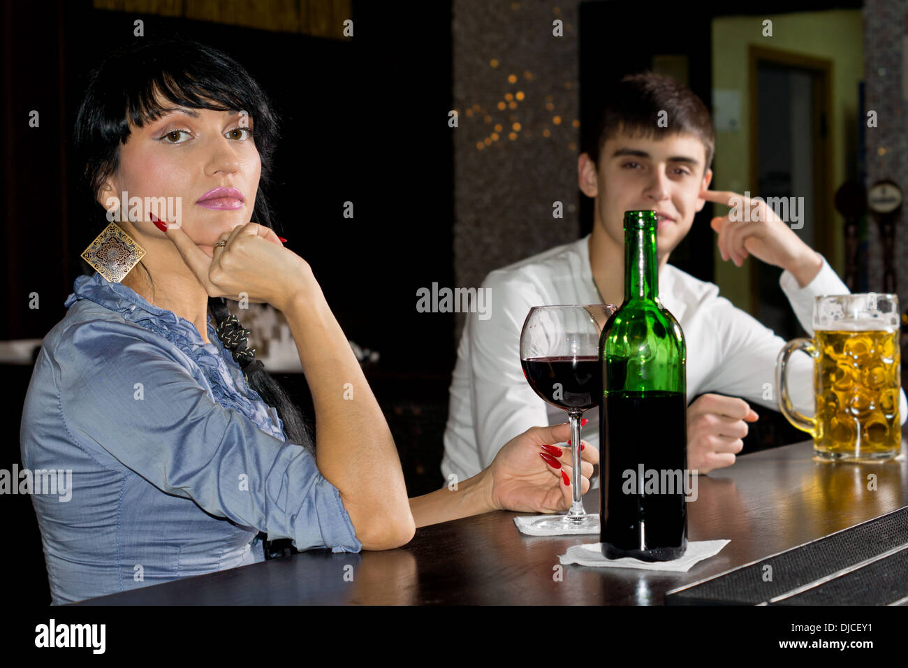 Sophisticated young woman drinking at a bar enjoying a bottle of red wine turning to look at the camera with a man in the background. Stock Photo