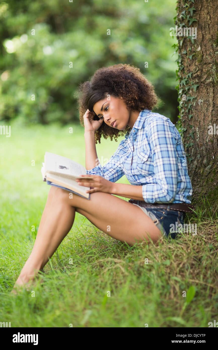 Gorgeous unsmiling brunette reading under tree Stock Photo