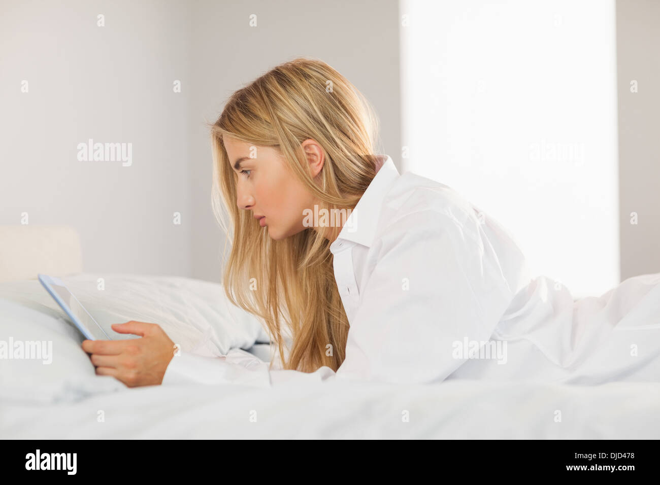 Pretty blonde wearing white mens shirt lying on bed using tablet Stock Photo