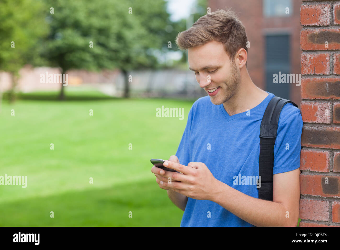 Smiling handsome student leaning against wall texting Stock Photo
