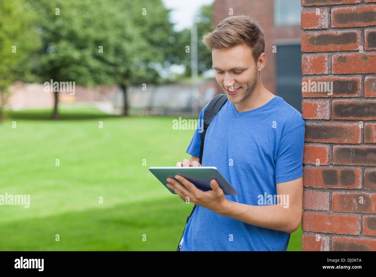 Smiling handsome student leaning against wall using tablet Stock Photo