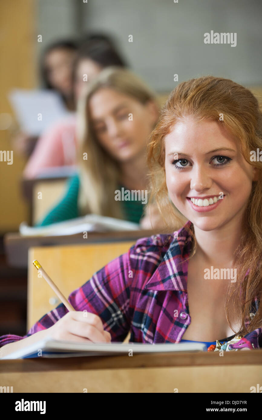 Smiling red head student taking notes in a lecture hall Stock Photo