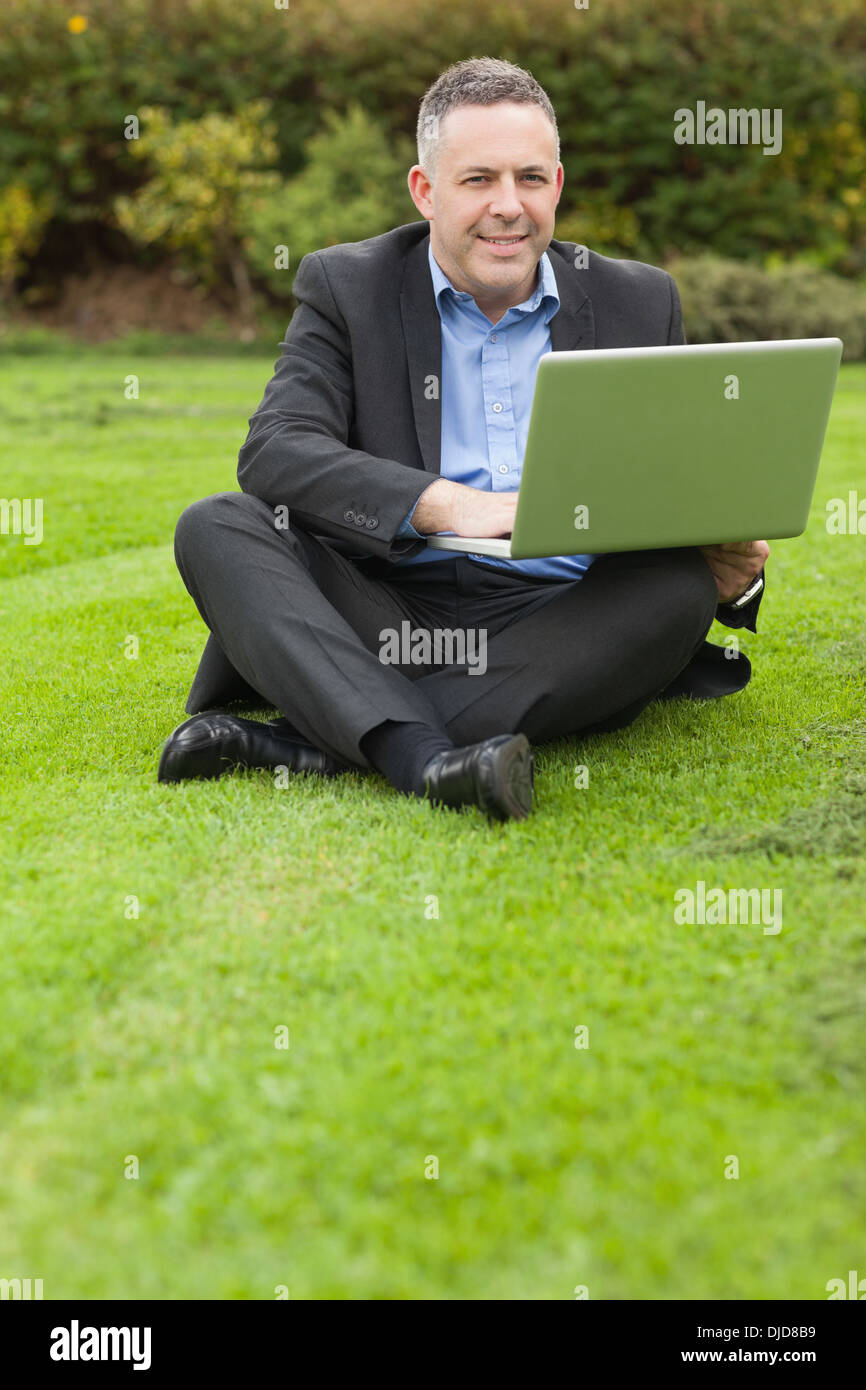 Happy lecturer sitting outside on campus using his laptop looking at camera Stock Photo