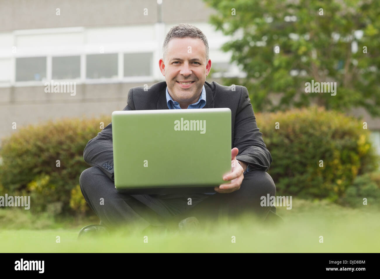 Smiling professor sitting outside on campus using his laptop looking at camera Stock Photo