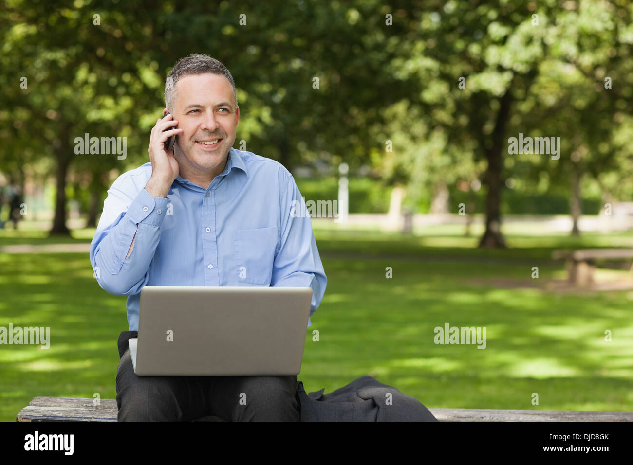 Happy professor talking on phone while sitting on bench using laptop on campus Stock Photo