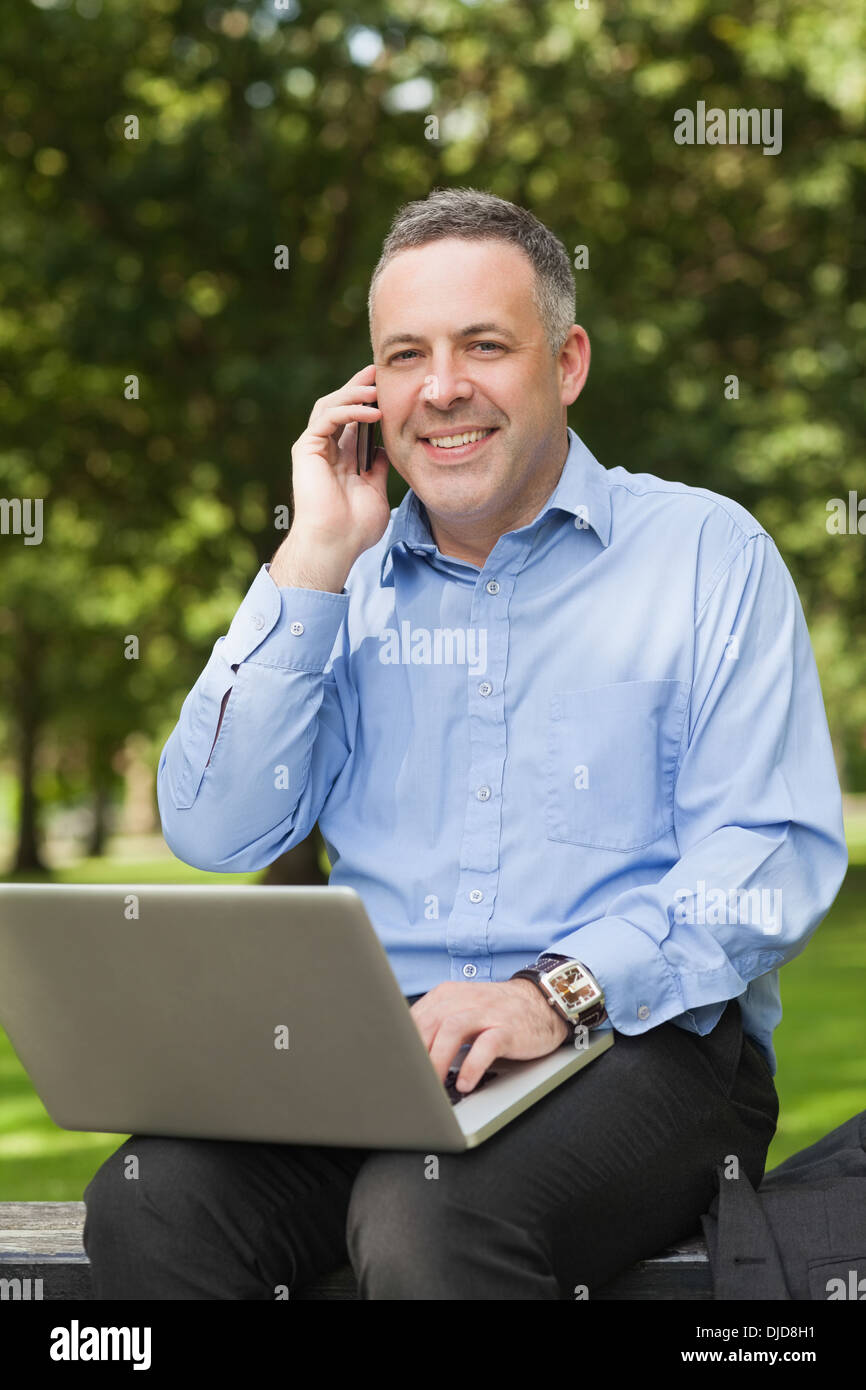 Laughing professor talking on phone while sitting on bench using laptop on campus Stock Photo