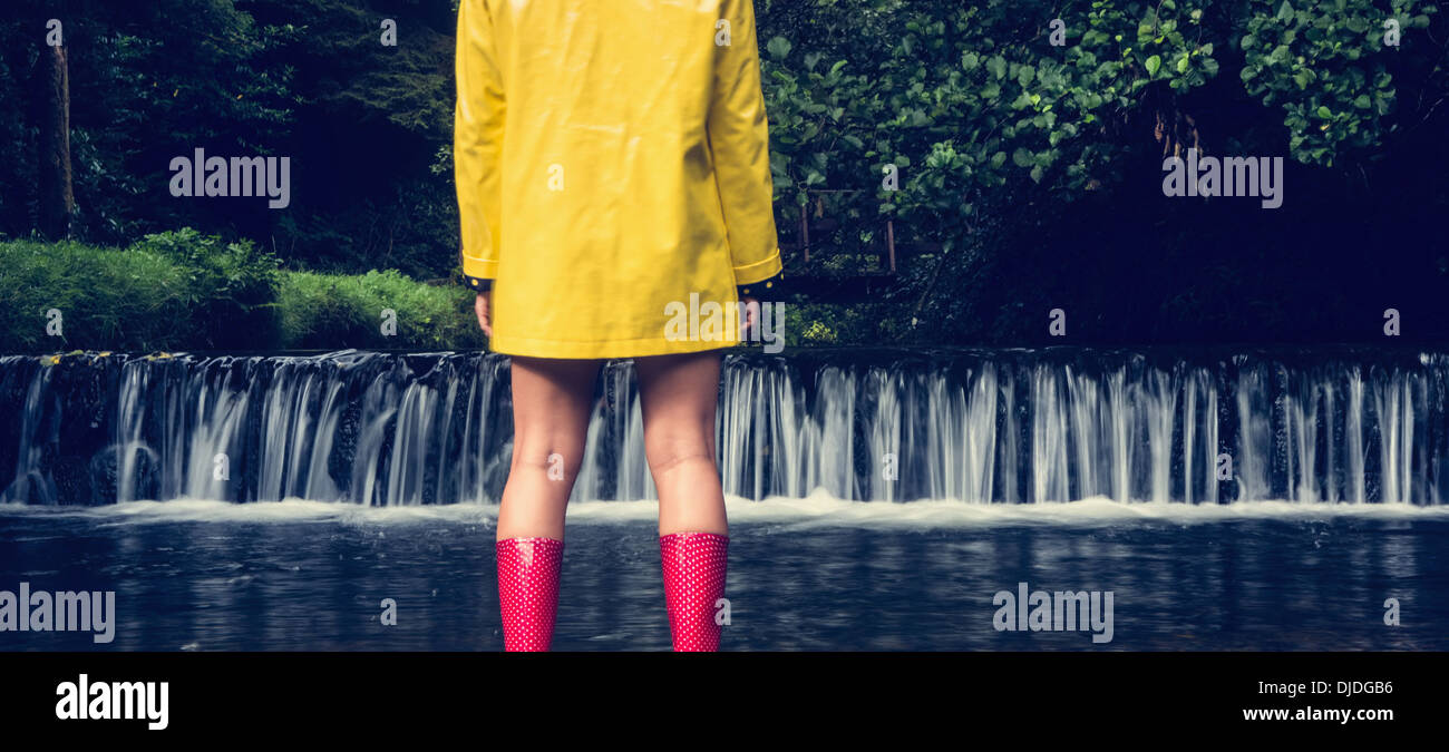 Woman in yellow raincoat and red gumboots looking at scenic waterfall Stock Photo