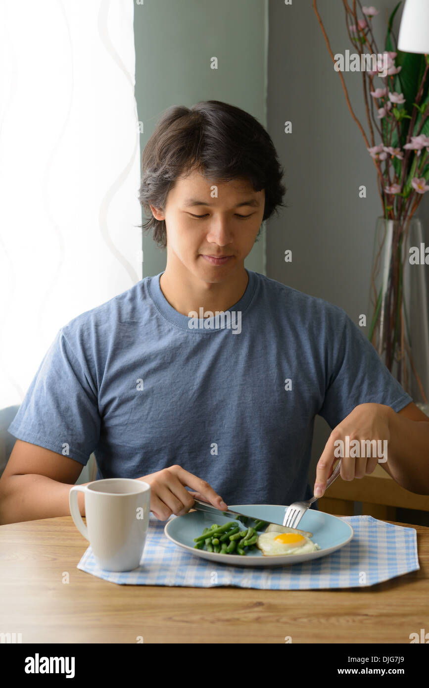 Young Asian man eating a healthy breakfast in his home, cutting egg, looking down, happy and content. Stock Photo