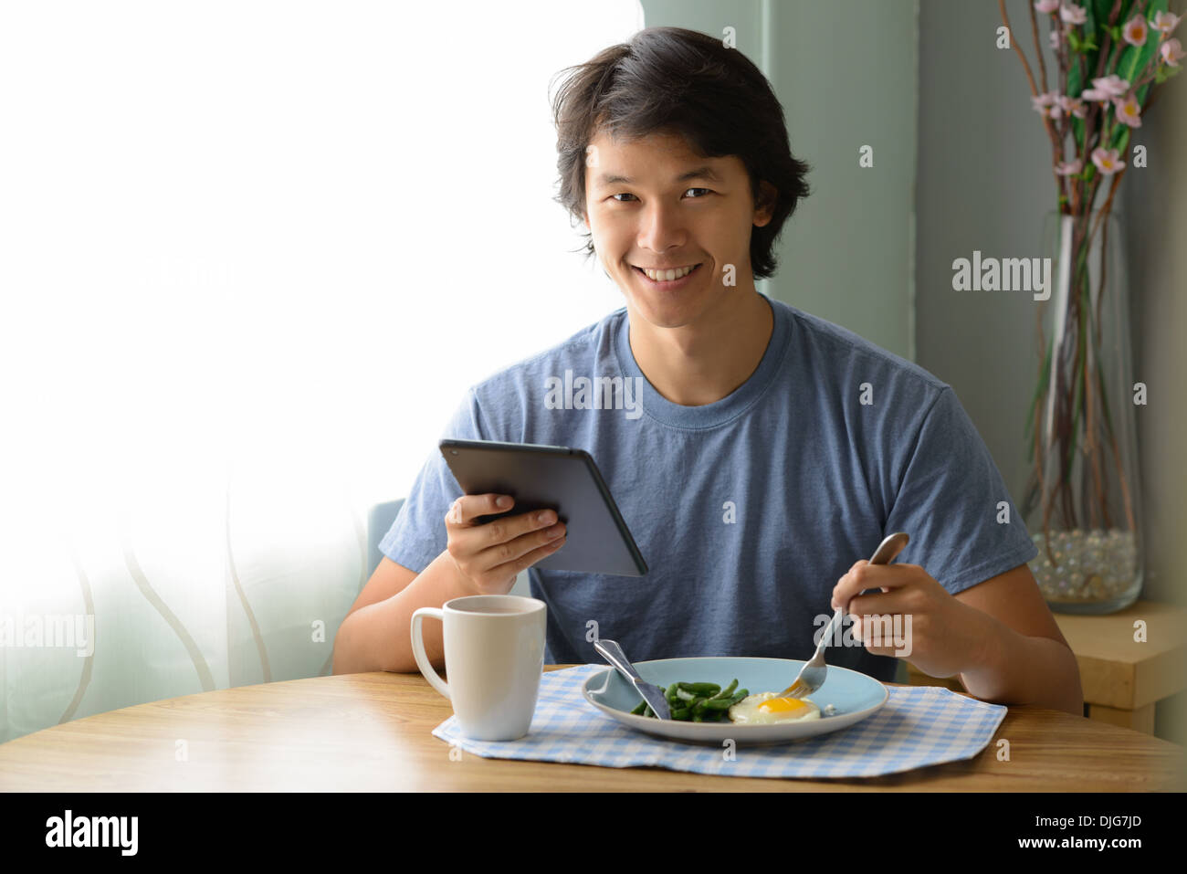 A young Asian man holding an ipad smiling and eating breakfast. Stock Photo