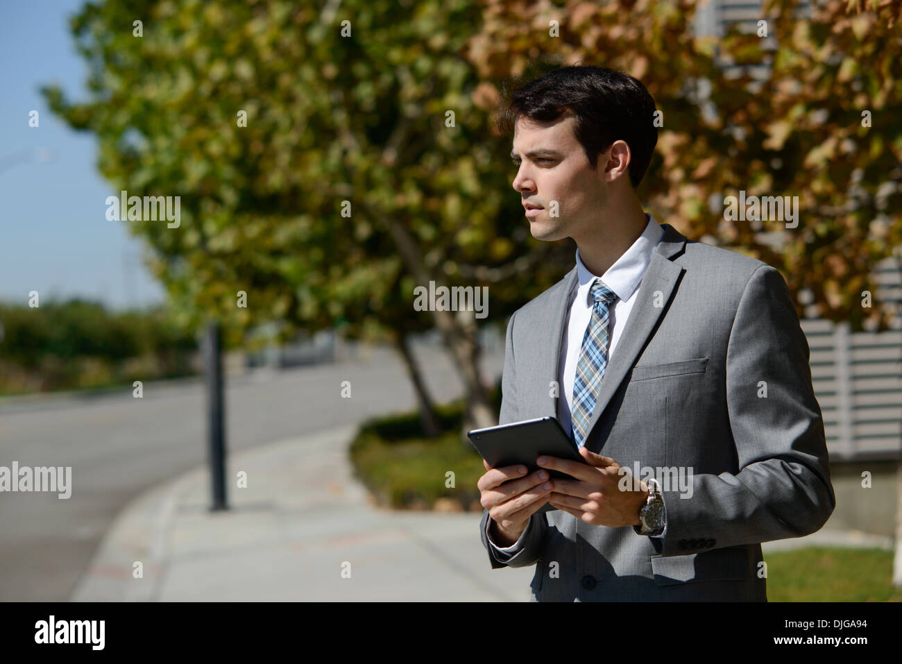A young business man holding an ipad, tablet, looking away ambitiously. He is standing on a footpath in front of a few trees. Stock Photo