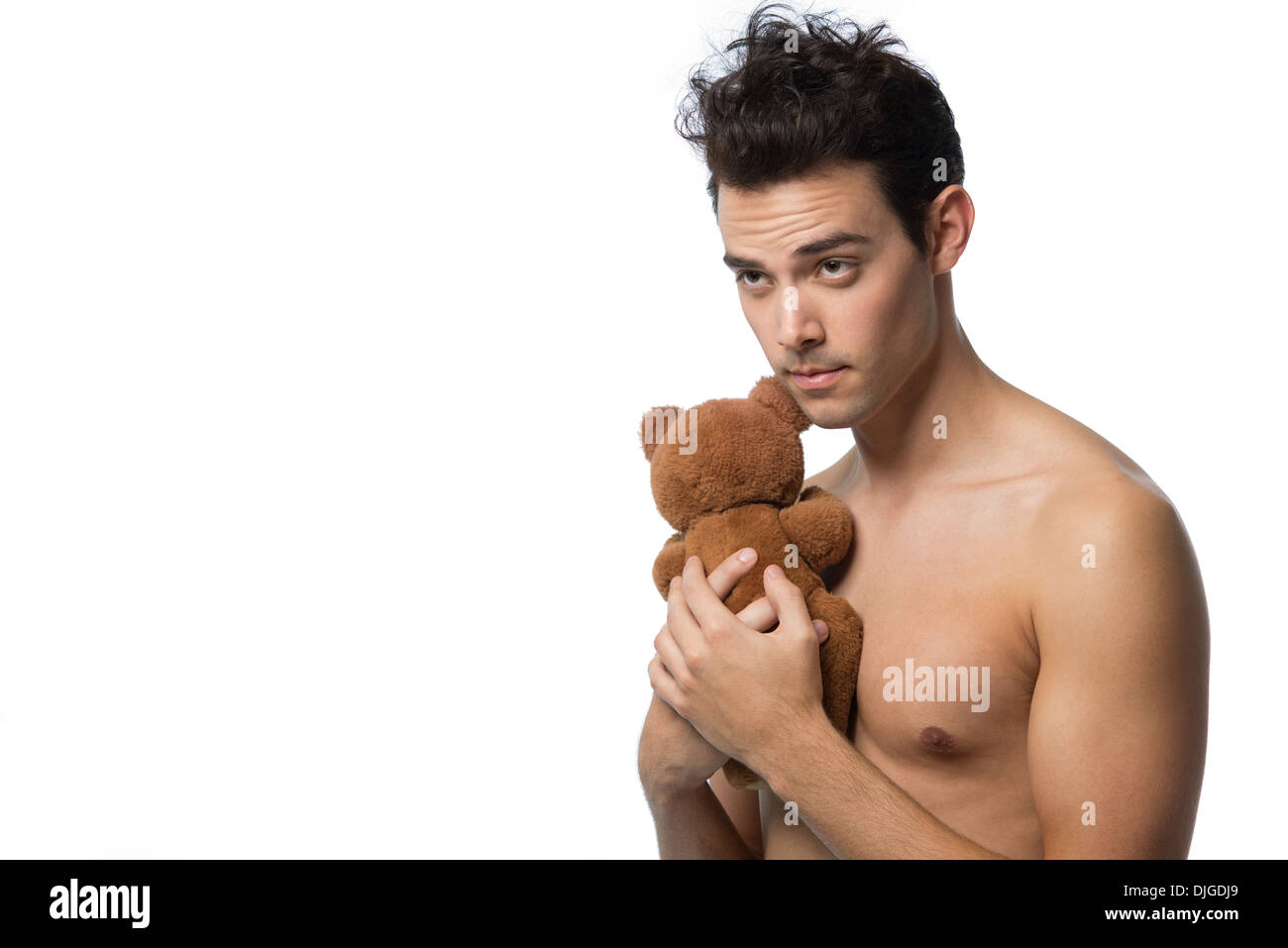 A dark hair handsome man, male model, bare chested, young at heart, hugging a brown teddy bear innocently, looking away happy. Stock Photo