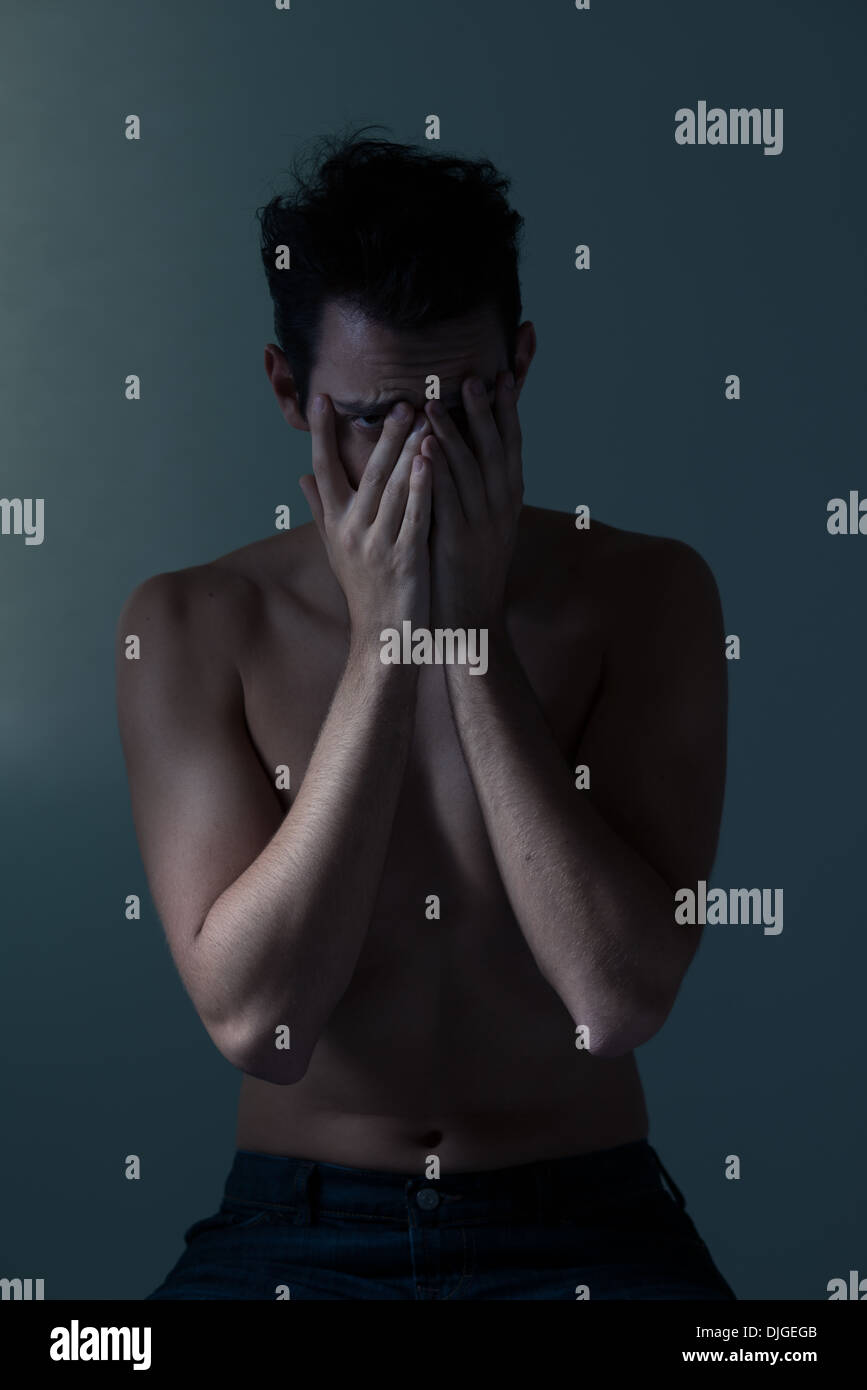 Caucasian young man sitting, covering face, looking at camera, shy and embarrassed. A conceptual moody photograph Stock Photo