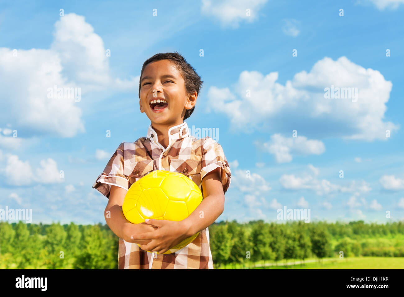 Laughing dark boy holding yellow volley ball standing in the park on sunny day with blue clouds on background Stock Photo