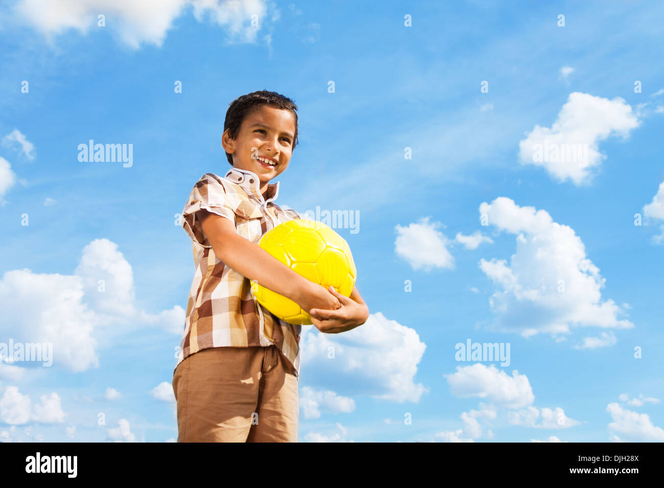 Cute little boy standing with football ball in hands over blue sky Stock Photo