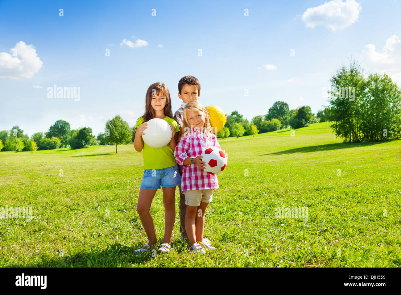 Three happy kids, boy and girls standing in the sunny summer park holding sport balls Stock Photo