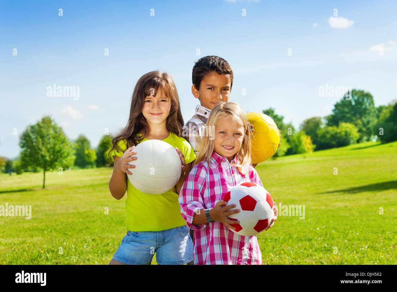 Team of three happy kids, boy and girls standing in the sunny summer park holding sport balls Stock Photo
