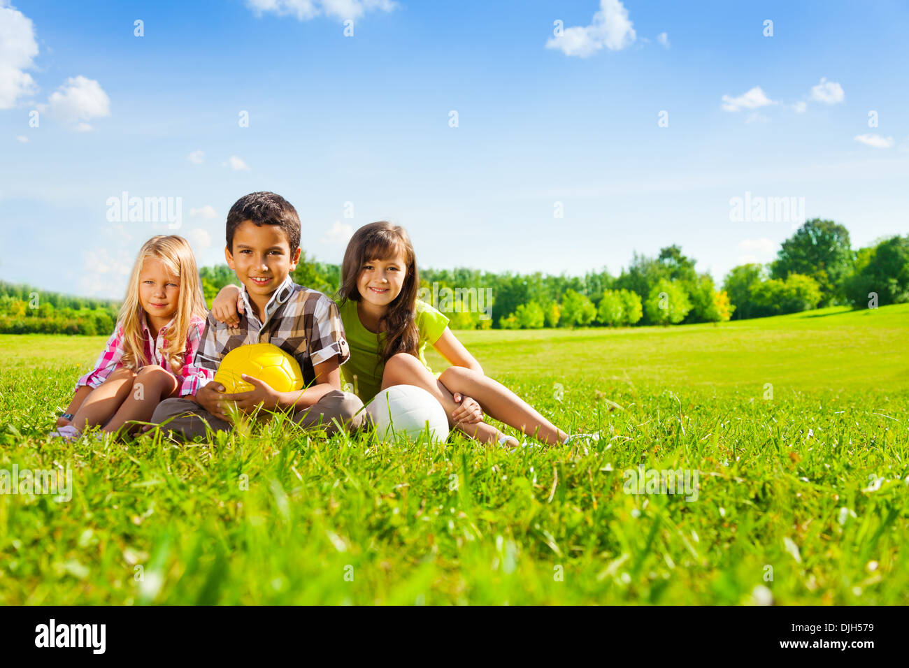 Three happy kids, boy and girls sitting in the sunny summer park holding sport balls Stock Photo