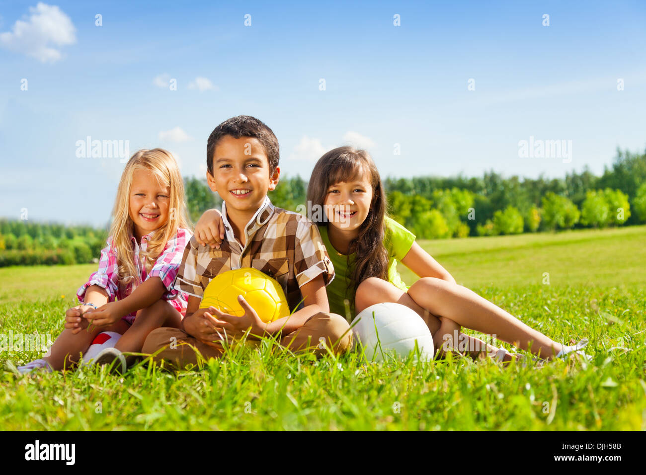 Portrait of three happy kids, boy and girls sitting in the sunny summer park holding sport balls Stock Photo