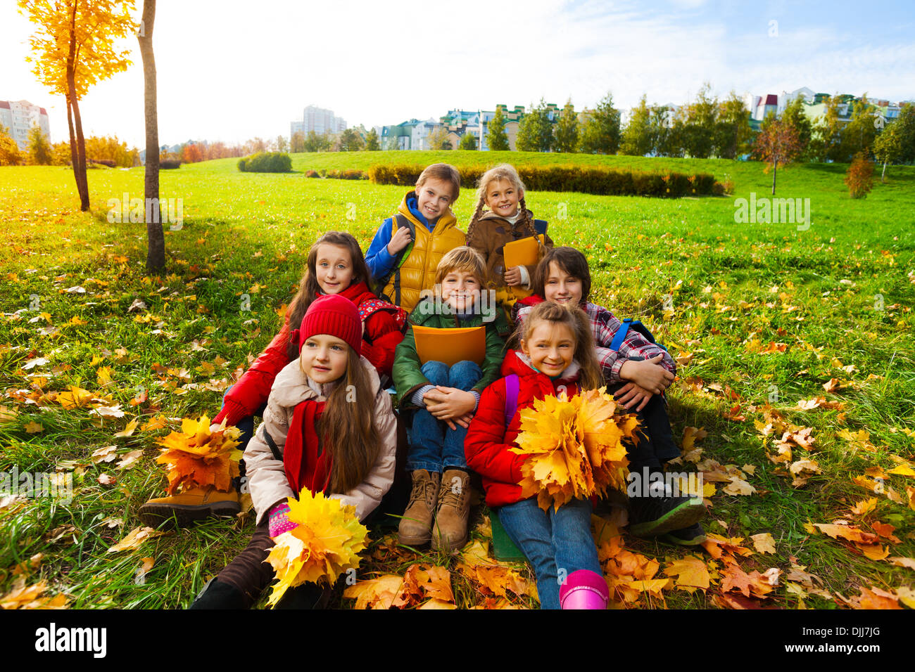 Group of happy kids sit in the park on the grass holding bouquets of autumn maple leaves and smiling Stock Photo