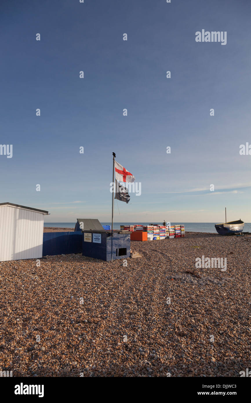 Flag of St George and the 'Jolly Roger' pirate flag fly on a shingle beach amidst fish containers and a fishing boat. Stock Photo
