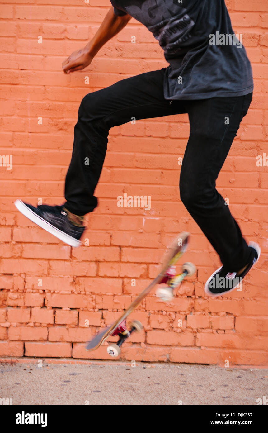 Young male in Deep Ellum performing a skateboarding trick called kickflip. Shot against a bright orange brick wall. Stock Photo