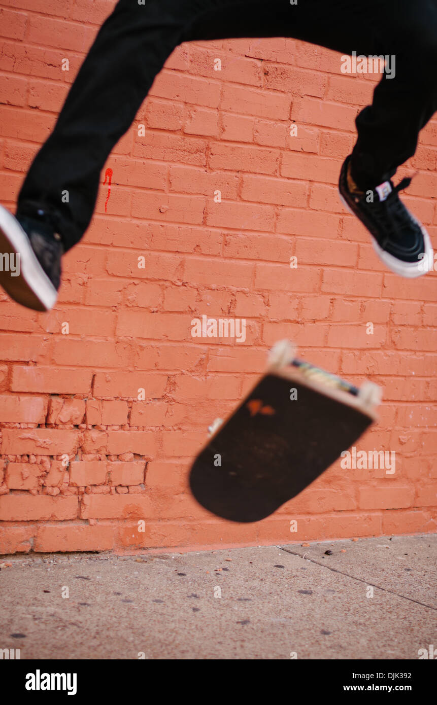 Young male in Deep Ellum performing a skateboarding trick called kickflip. Shot against a bright orange brick wall. Stock Photo