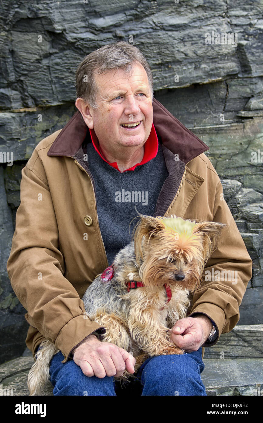 Mature man, sitting near cliffs on a beach, with his pet Yorkshire terrier dog on his lap, Harlyn Bay, Padstow, Cornwall, Englan Stock Photo
