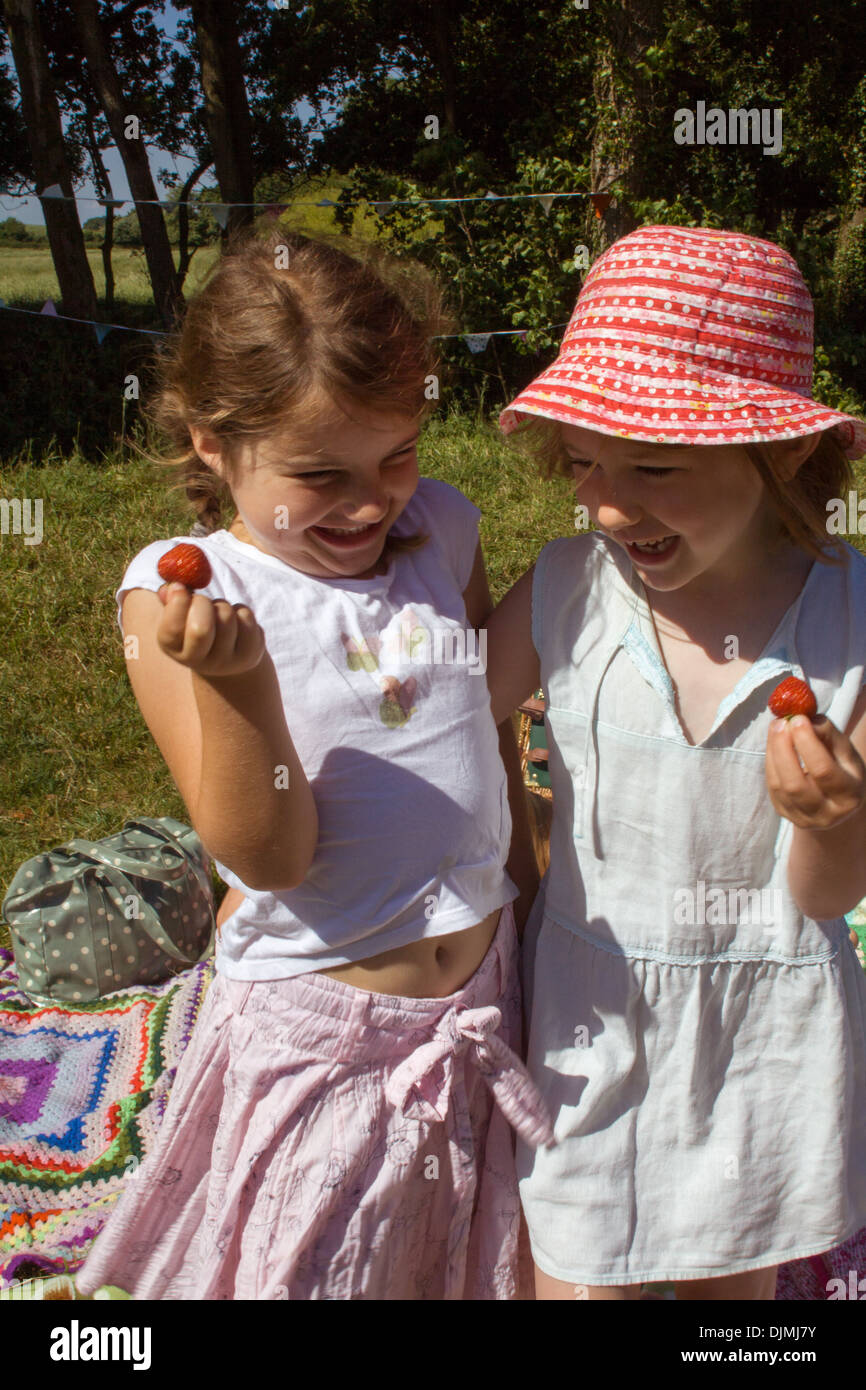 Two young girls laughing and having fun out in the sunshine on a picnic, holding strawberries and having fun in Somerset, UK. Stock Photo