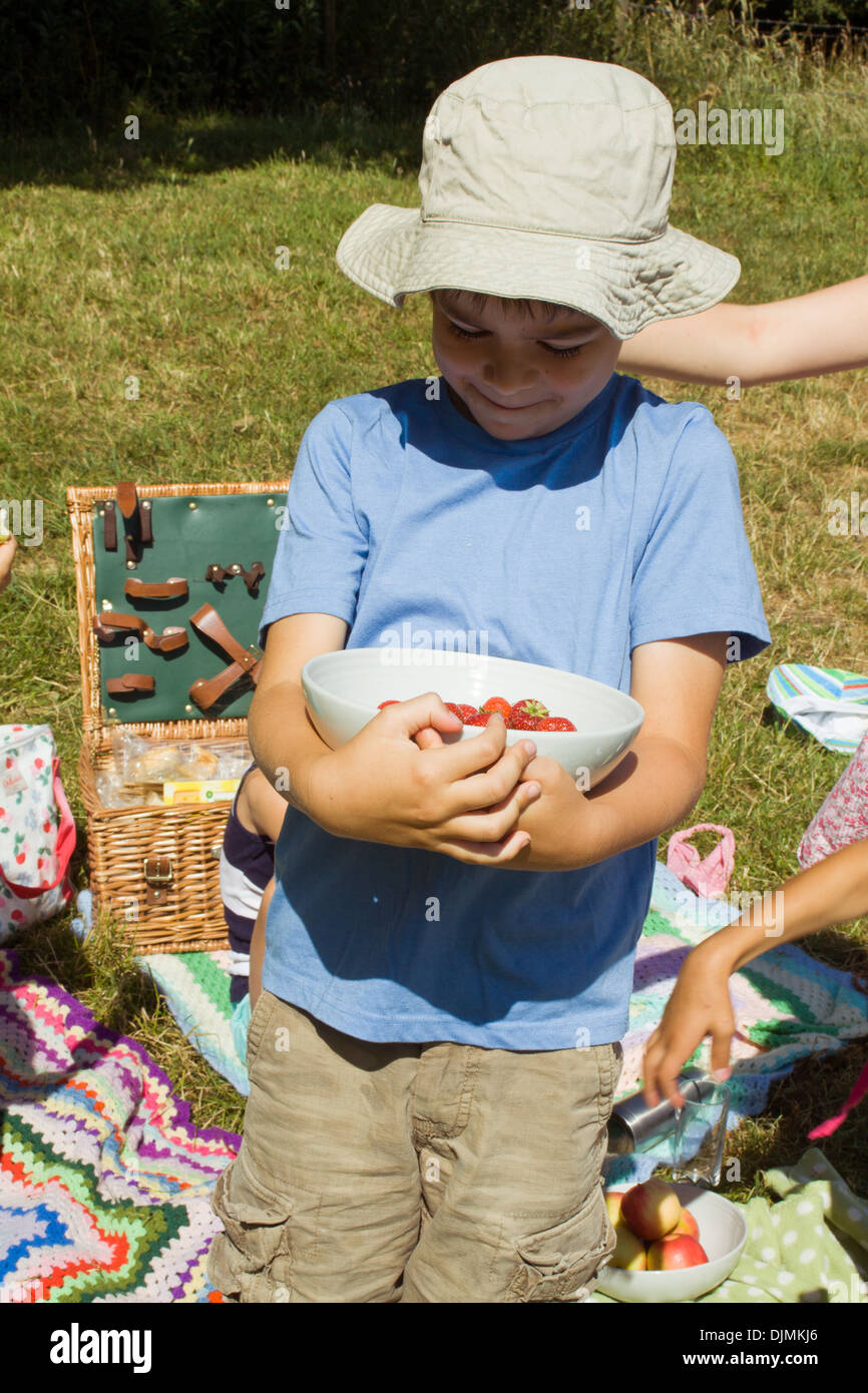 A young boy wearing a blue t-shirt and a white sun hat holding and looking at a bowl of strawberries in Somerset, UK. Stock Photo