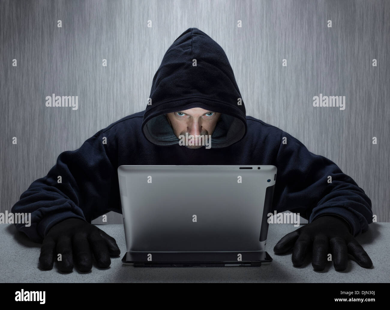 A hooded male representing a cyber criminal, looking at a tablet computer. Stock Photo