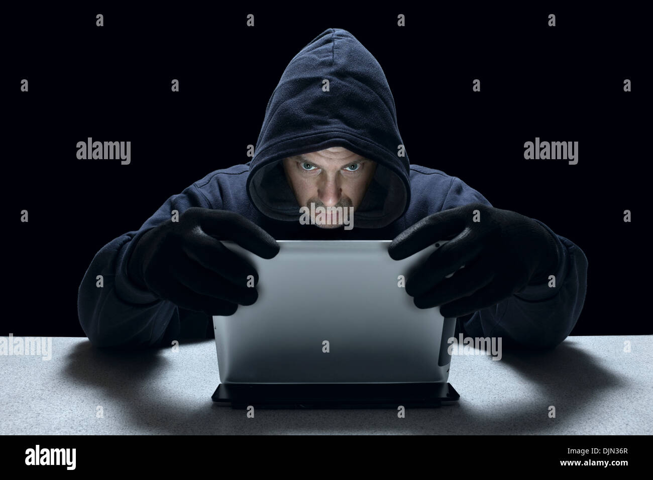 A hooded male representing a cyber criminal, using a tablet computer. Stock Photo