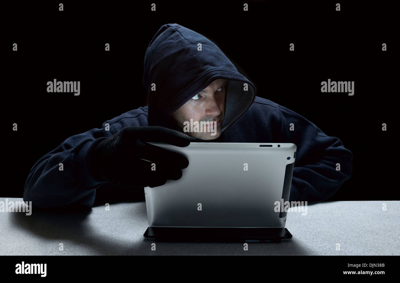 A hooded male representing a cyber criminal, using a tablet computer. Stock Photo