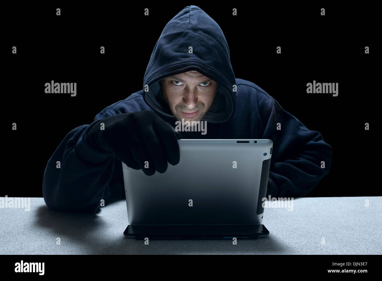 A hooded man using a tablet computer representing a cyber criminal. Stock Photo