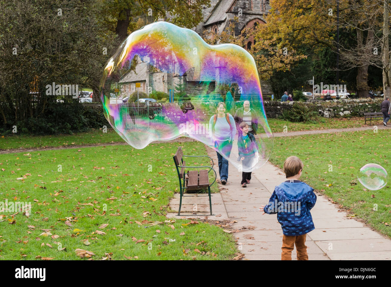 A young boy has fun with a giant soap bubble showing rainbow colours and people smiling in Betws-y-Coed, Conwy, North Wales, UK Stock Photo