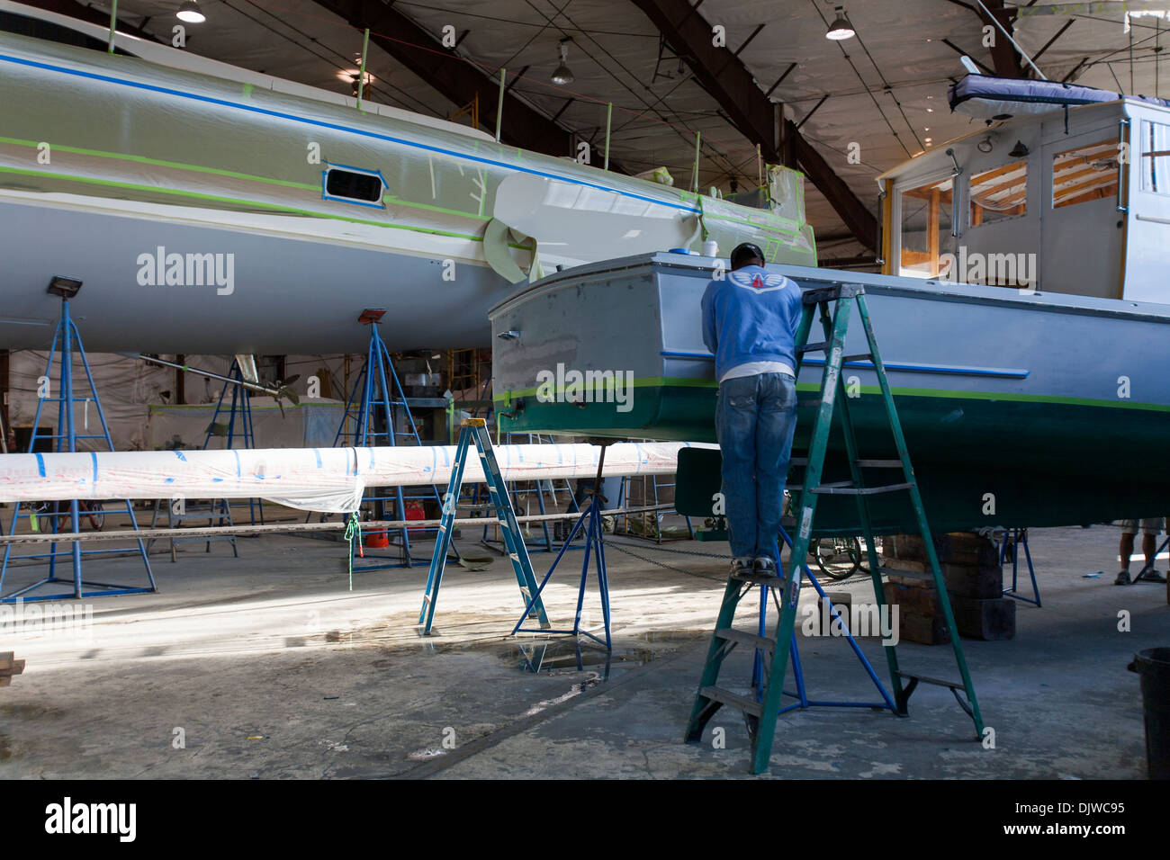 A painter works of a wooden lobster boat while standing on a ladder in a large work shed at Newport Shipyard in Rhode Island Stock Photo