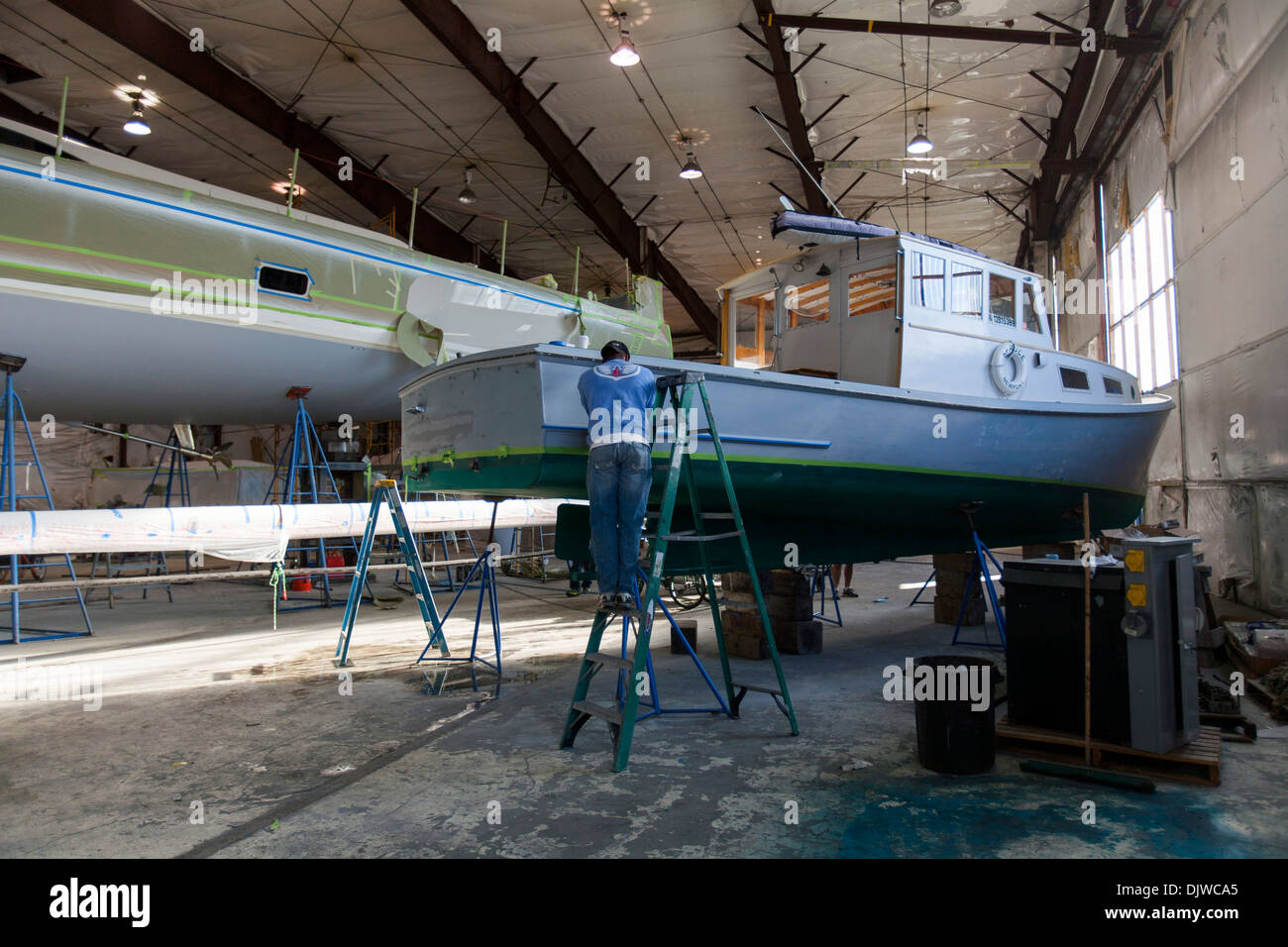 A painter works of a wooden lobster boat while standing on a ladder in a large work shed at Newport Shipyard in Rhode Island Stock Photo