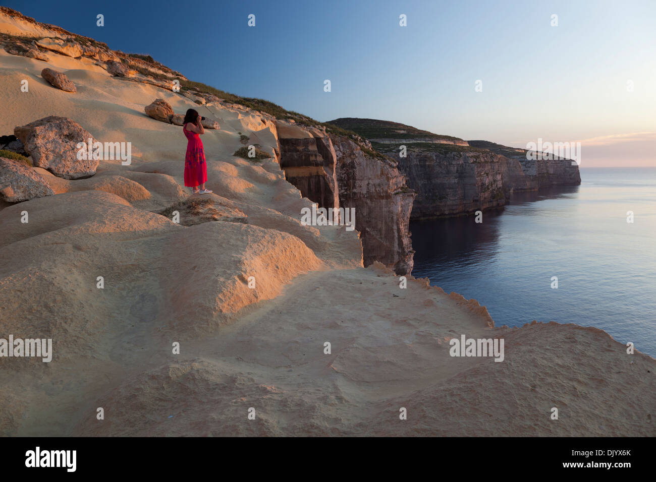A woman taking pictures of the setting sun from atop the seacliffs at the northwesterly coast of Gozo. Stock Photo