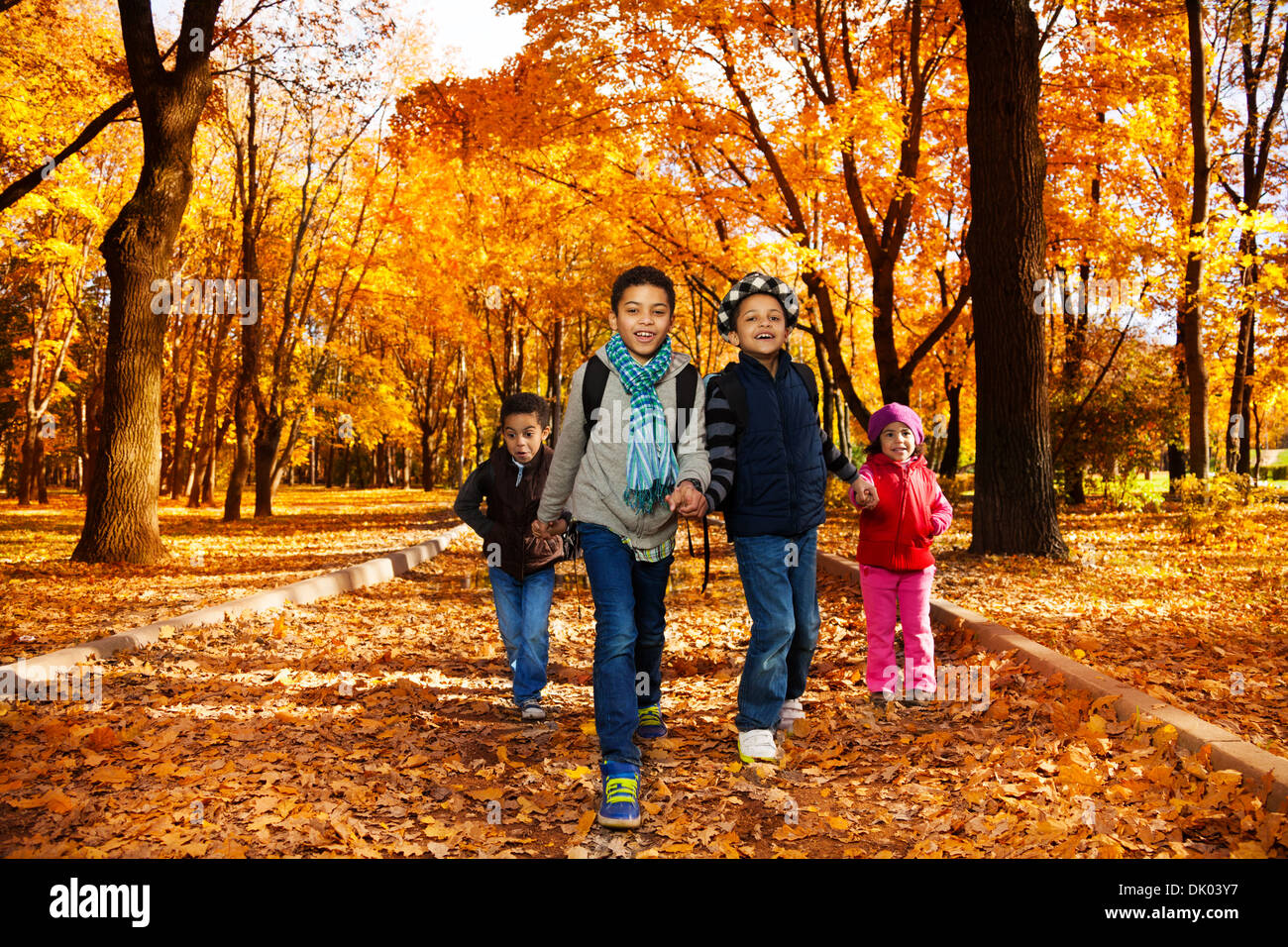 Group of four black boys and girl, happy brothers and sister 3-10 years old going together holding hands in the park wearing backpacks and autumn clothes in maple park Stock Photo