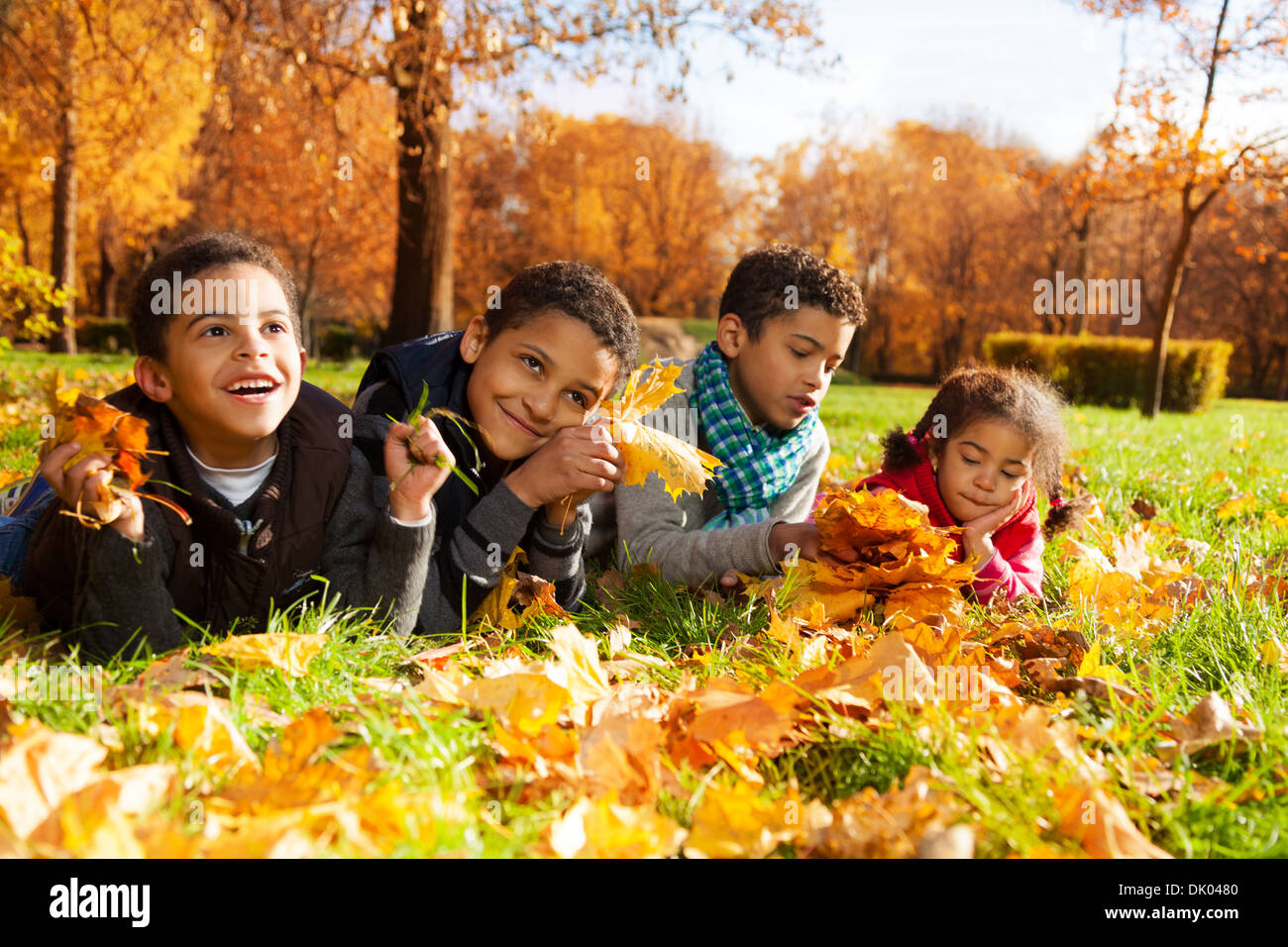 Group of four black boys and girl, happy brothers and sister 3-10 years old laying in grass together in the park in autumn clothes under maple park with orange leaves Stock Photo