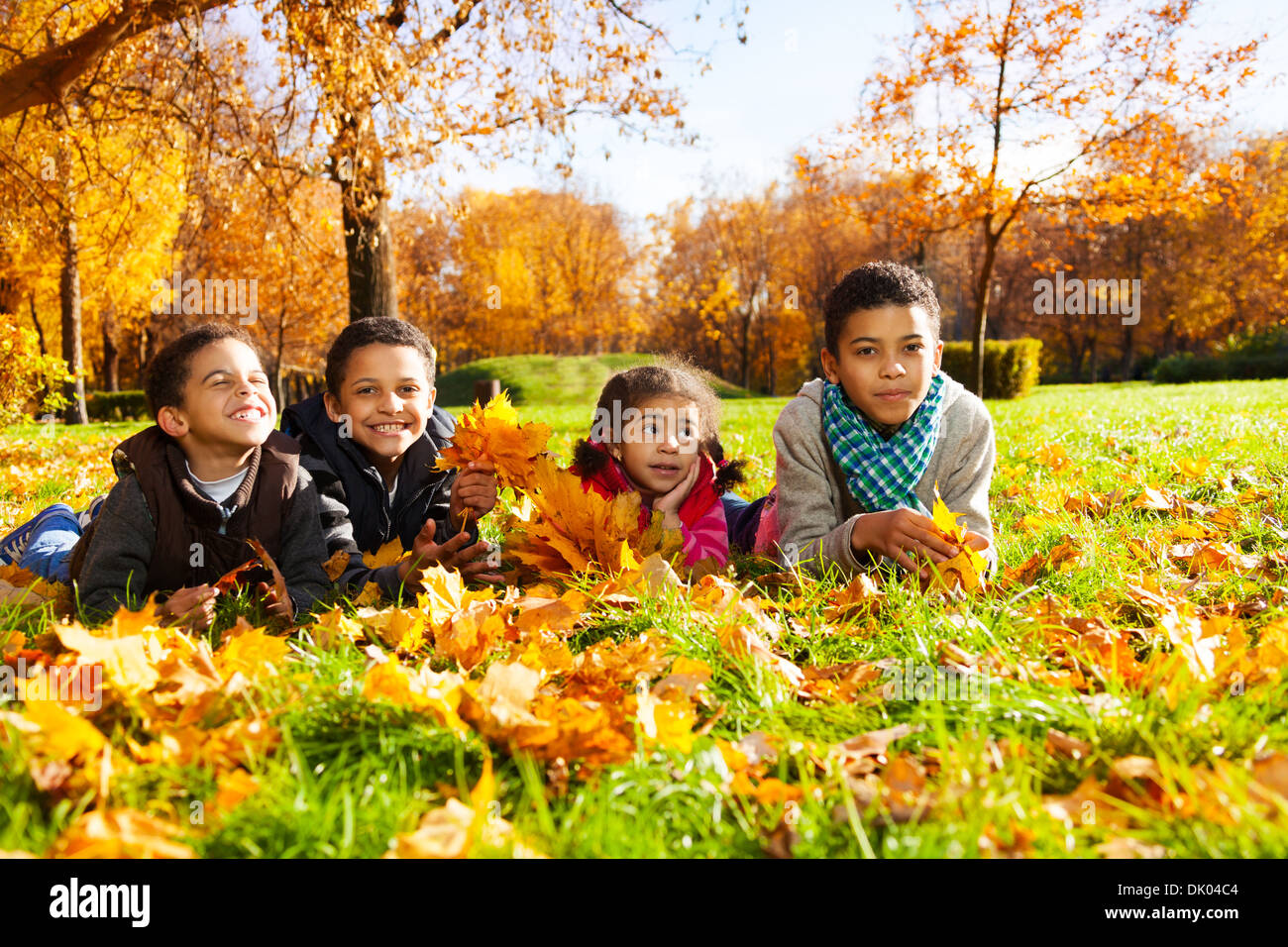 Group of four black boys and girl, happy brothers and sister 3-10 years old laying in grass together in the park in autumn clothes holding maple leaves bouquets Stock Photo