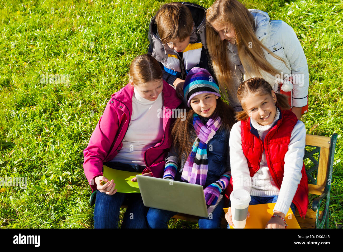 Close portrait of a group of five school kids, boy and girls sitting on the bench in autumn park with laptop at sunny day, happy and smiling Stock Photo