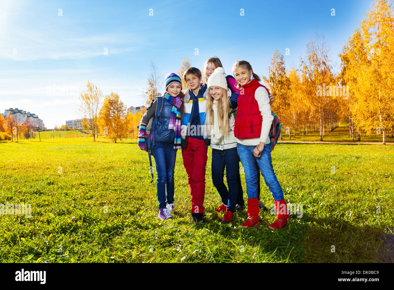 Group of five happy kids, boys and girls standing in the park after school wearing warm clothes with backpacks on sunny autumn day Stock Photo