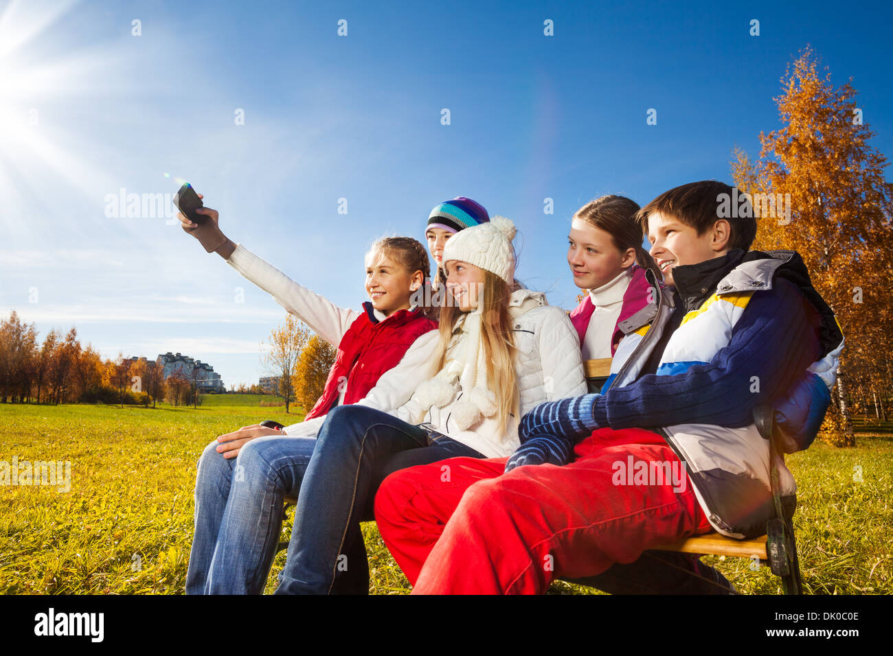 Group of kids sitting on the bench and self photographing with camera phone, boy and girls on autumn sunny day Stock Photo