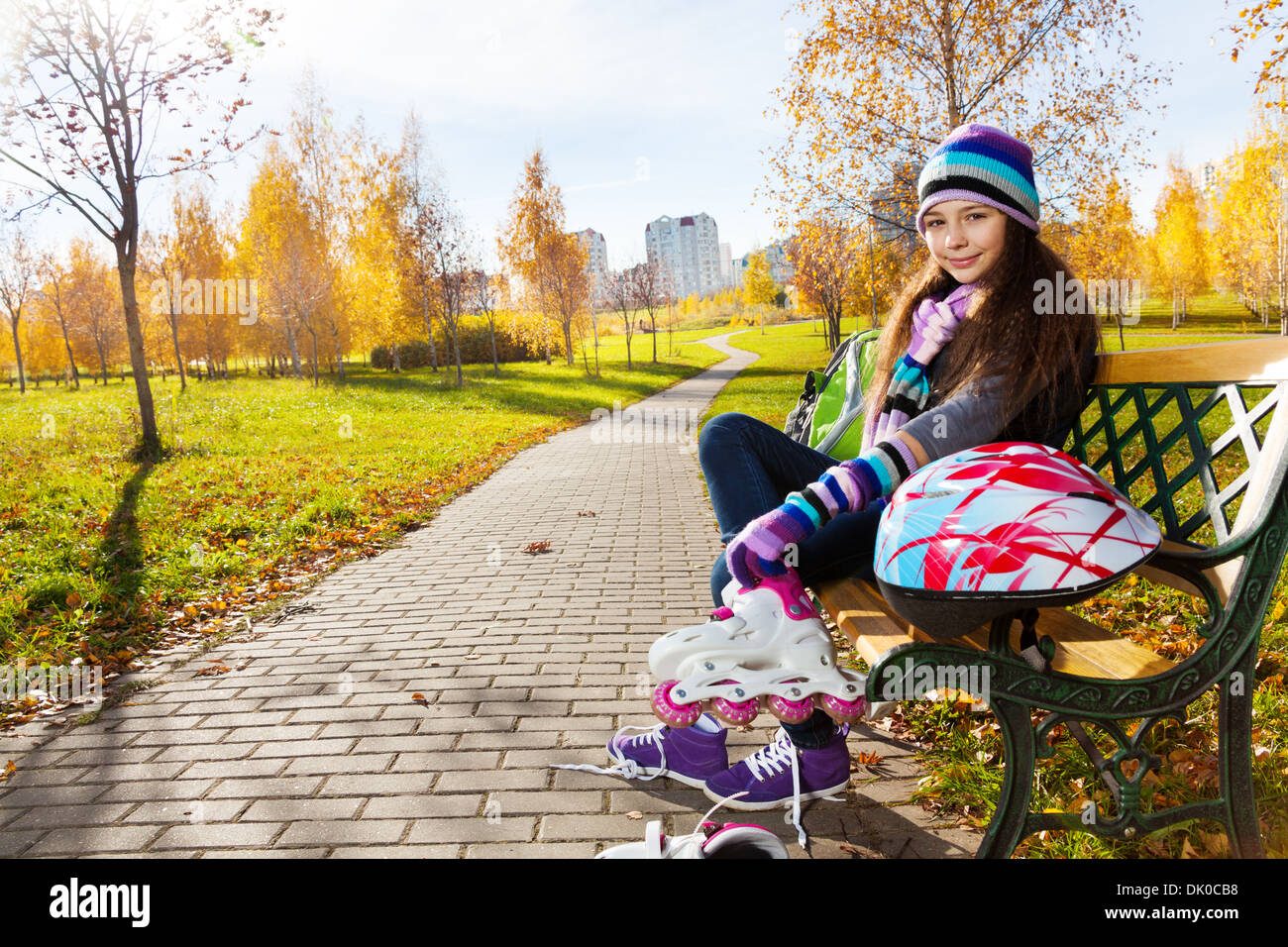 11 years old beautiful girl sitting on the bench in the park putting on roller blades to go skating in the autumn park on sunny day Stock Photo